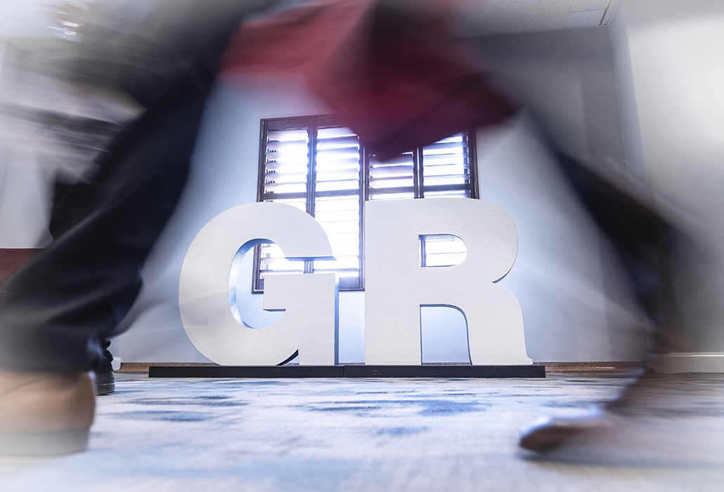 Large white letters 'GR' in a blurred indoor setting with people walking past them.