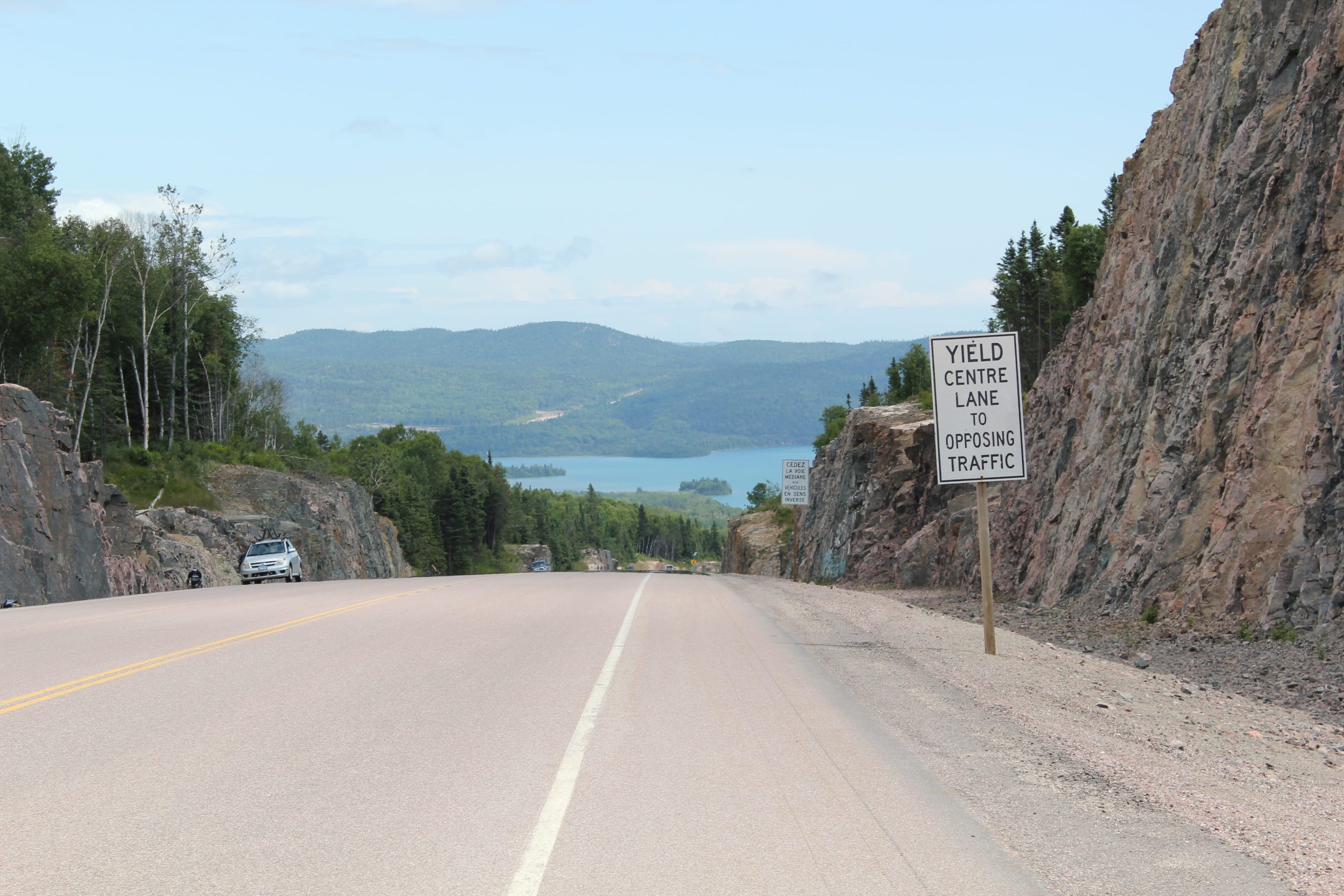 A scenic road with a yield sign indicating to yield to opposing traffic, surrounded by rocky cliffs and a lake in the background.