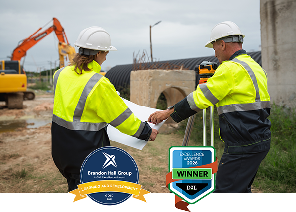 Two people in hard hats reviewing construction plans at a work site, with award logos displayed in the foreground.